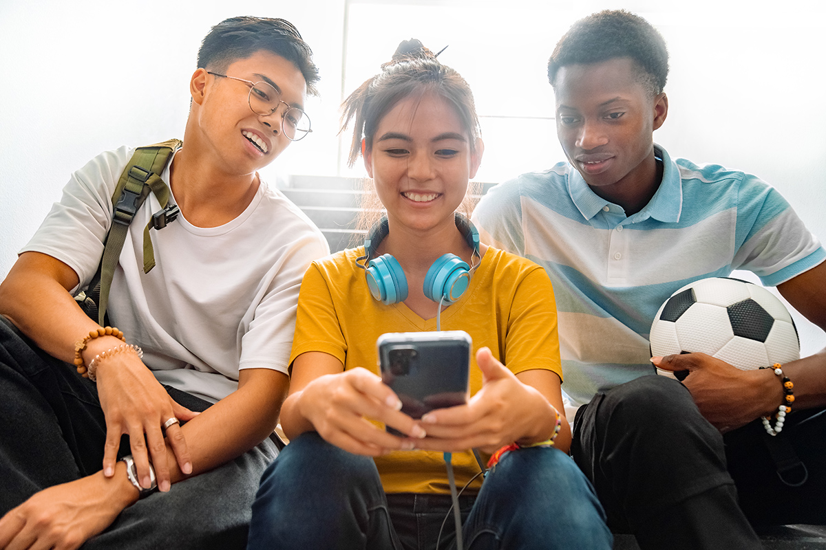 Three teens – two males and one female – sit together in a stair well. They are looking at a phone that the female is holding.