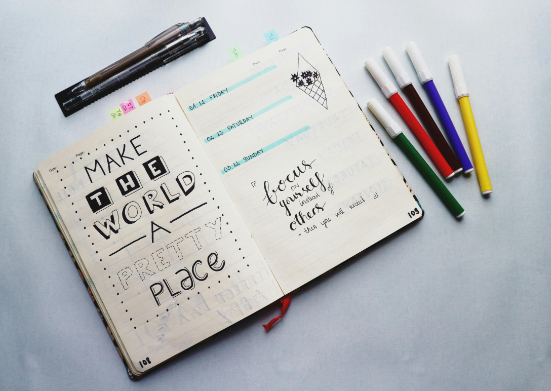 Jot journal open on a white counter to a page that says "make the world a better place" with coloured markers lying to the side