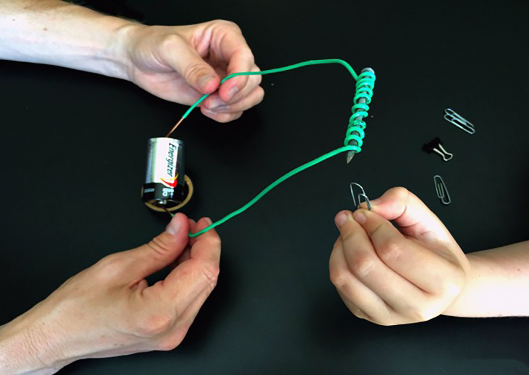 Green wire wrapped around a nail, an adult's hands holding the bare ends of the wire against each end of a battery, and a child's hand holding a paperclip up to the end of the nail.