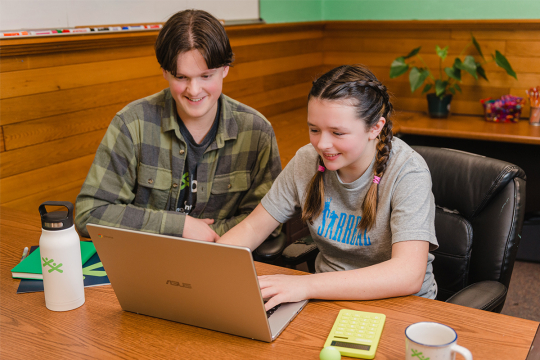 Two youth smiling and looking at a laptop on a desk.