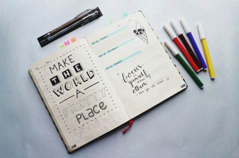 Jot journal open on a white counter to a page that says "make the world a better place" with coloured markers lying to the side