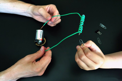 Green wire wrapped around a nail, an adult's hands holding the bare ends of the wire against each end of a battery, and a child's hand holding a paperclip up to the end of the nail. 