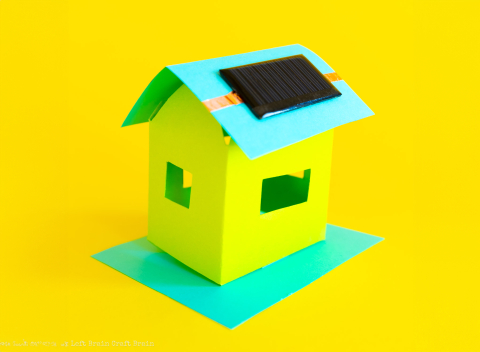 Golden yellow background with a small simple house crafted from bright yellow and aqua paper, with a small solar panel attached to the roof.